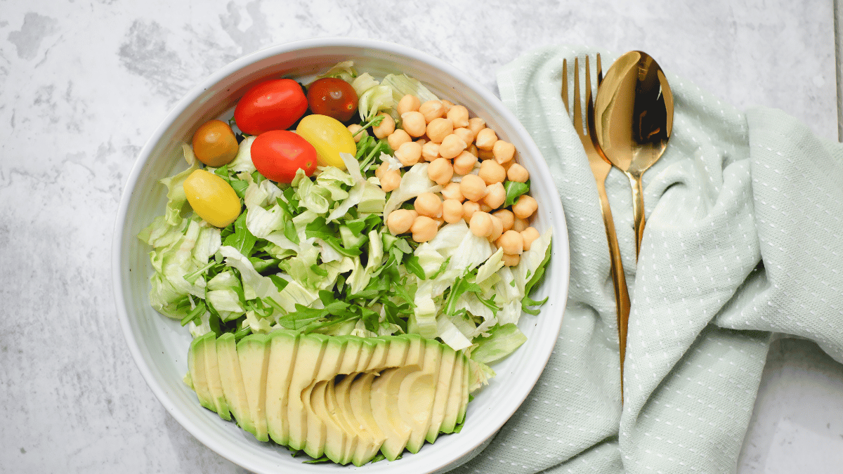 Tigela contendo uma salada fresca com alface, tomates cereja coloridos, grão-de-bico e fatias de abacate, acompanhada por talheres dourados e um guardanapo verde claro.