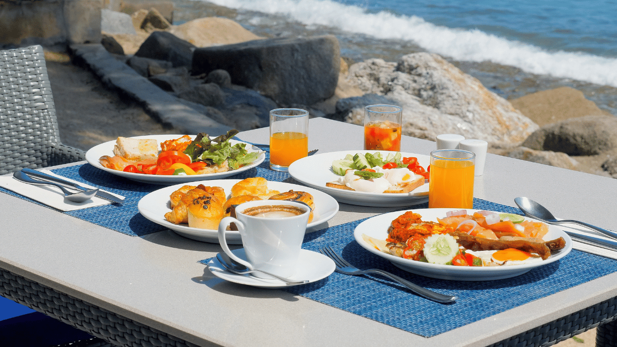 Mesa ao ar livre com pratos variados de café da manhã, incluindo salada, frutas, pães, sucos e café, com vista para o mar ao fundo.