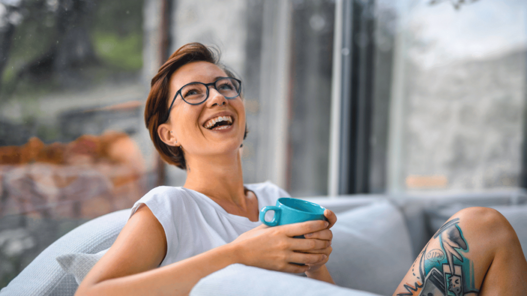  Mulher sorridente usando óculos, segurando uma caneca azul enquanto relaxa em um sofá, com um ambiente iluminado e moderno ao fundo.