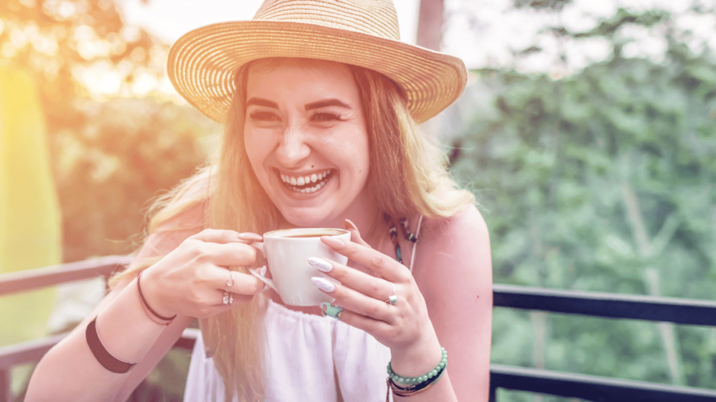 Mulher sorridente segurando uma xícara de café ao ar livre, usando um chapéu de palha, com fundo verde de natureza.