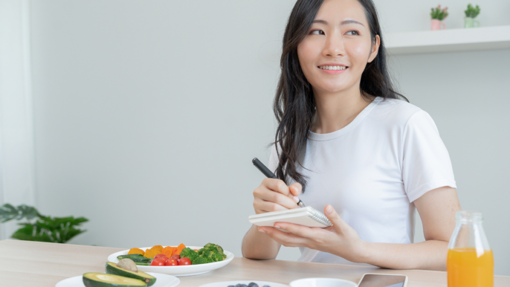 Mulher sorridente com um bloco de notas nas mãos, sentada em uma mesa com alimentos saudáveis, incluindo vegetais e frutas, representando planejamento alimentar.