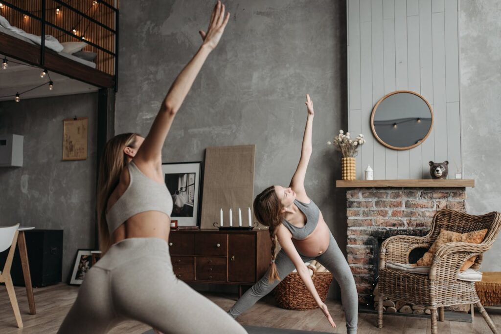 Duas mulheres praticando yoga em um ambiente interno aconchegante, com decoração rústica, espelho redondo na parede e móveis de madeira.