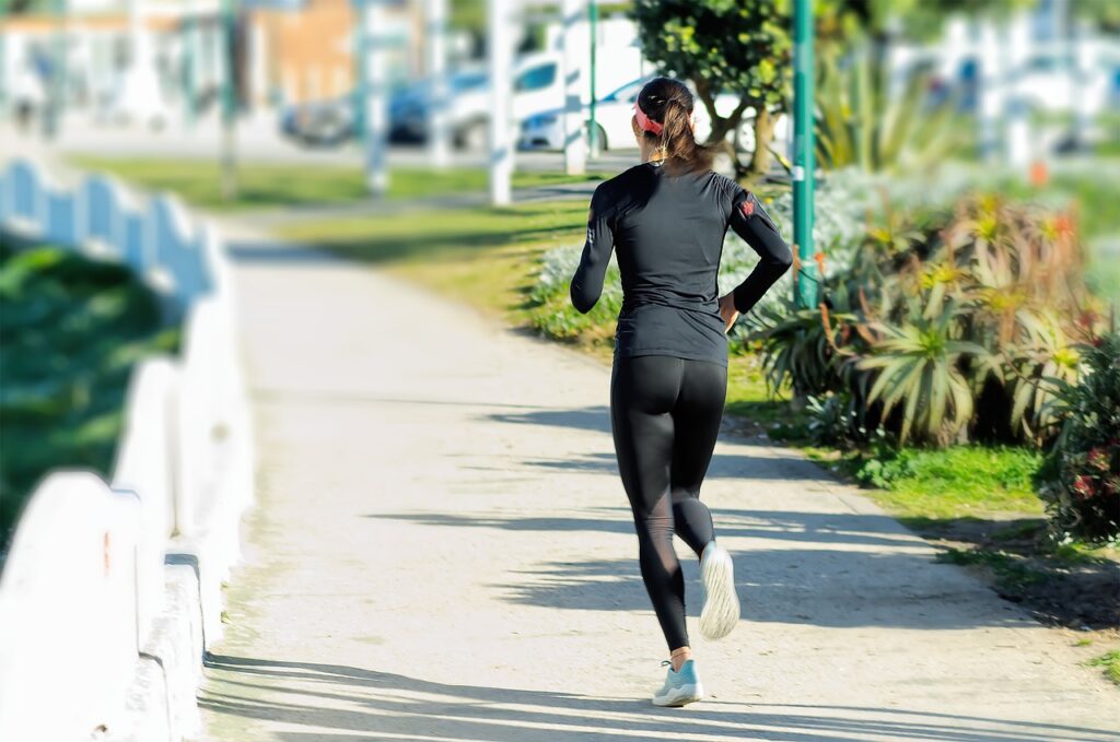 Mulher correndo em uma trilha ao ar livre em um ambiente urbano com vegetação ao redor, vestindo roupa esportiva preta.