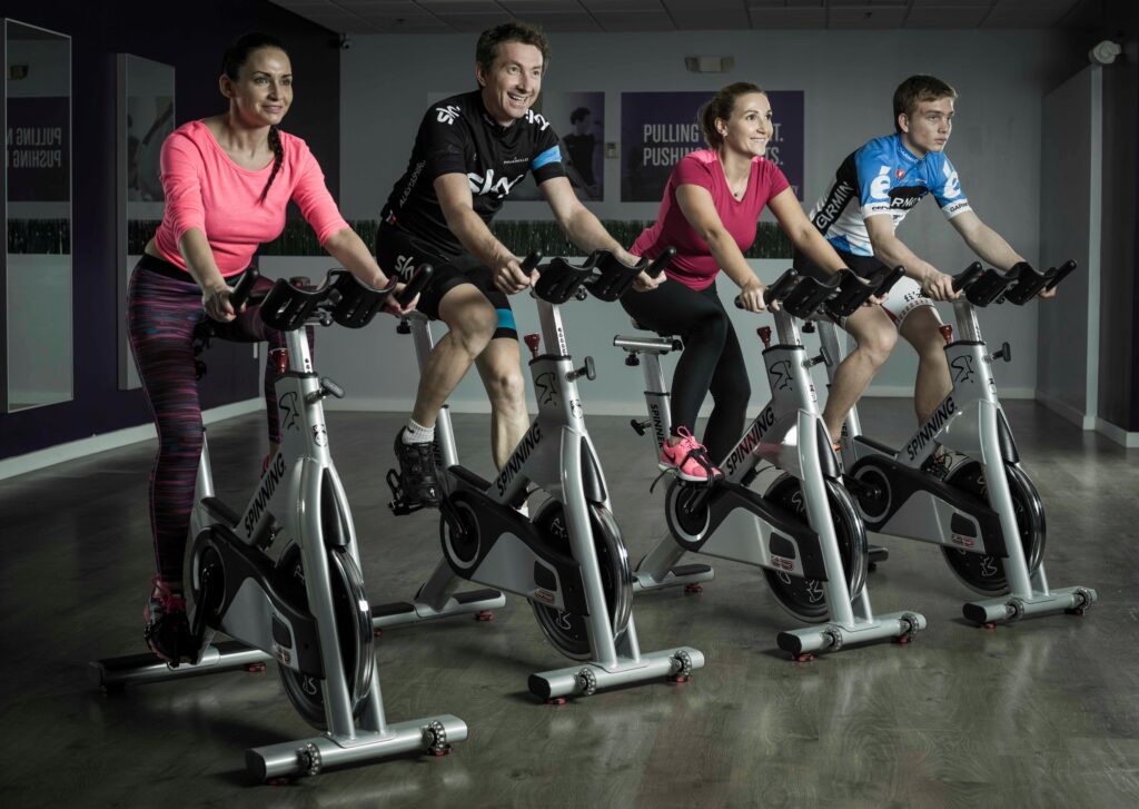  Quatro pessoas pedalando em bicicletas de spinning em uma aula de grupo, sorrindo e se exercitando em uma academia.