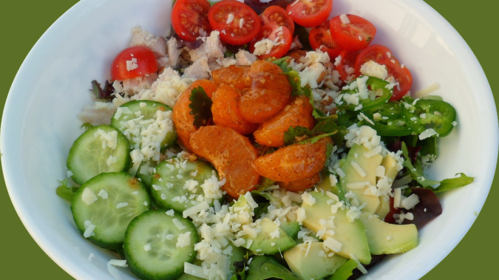 Uma salada fresca e colorida composta por tomates-cereja, pepino, abacate, tangerinas, queijo ralado e pedaços de frango desfiado, com folhas verdes e pimenta verde. A salada está em um recipiente branco, pronta para ser servida como uma refeição saudável.