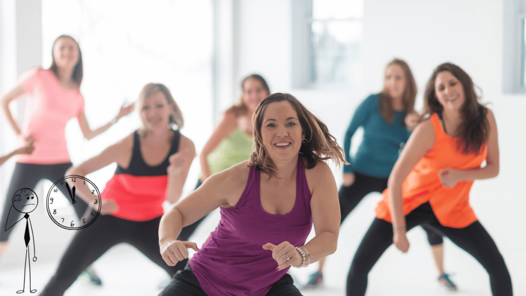 Grupo de mulheres se exercitando em uma aula de dança fitness, com uma delas sorrindo e com roupas coloridas, enquanto um desenho de relógio e um personagem pensativo são exibidos na parte inferior esquerda da imagem.