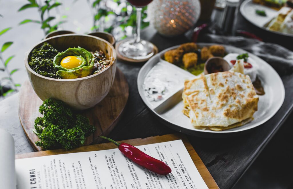 Uma refeição saudável composta por uma tigela de quinoa com abacate, verduras e um ovo cru, acompanhada de uma porção de pão sírio com molho e falafel. No fundo, há um cardápio de restaurante e uma pimenta vermelha ao lado.