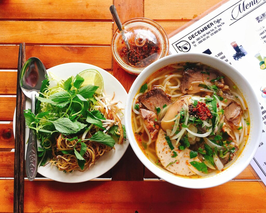 Um prato de pho, uma sopa vietnamita com macarrão, carne de boi, cebolas e ervas frescas, acompanhado de uma porção de brotos de feijão, limão e folhas de hortelã.