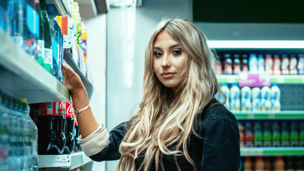 Mulher loira com cabelos longos e ondulados está em um corredor de supermercado, observando a prateleira de bebidas.