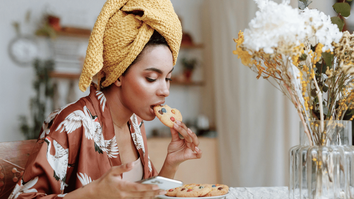 Mulher com toalha na cabeça e roupão estampado saboreia um biscoito com gotas de chocolate enquanto usa o celular, sentada à mesa com flores decorativas ao fundo.