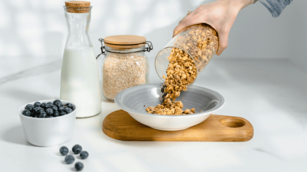 Uma mão despeja granola em um prato, acompanhado por leite, aveia e mirtilos, compondo um café da manhã saudável e equilibrado.