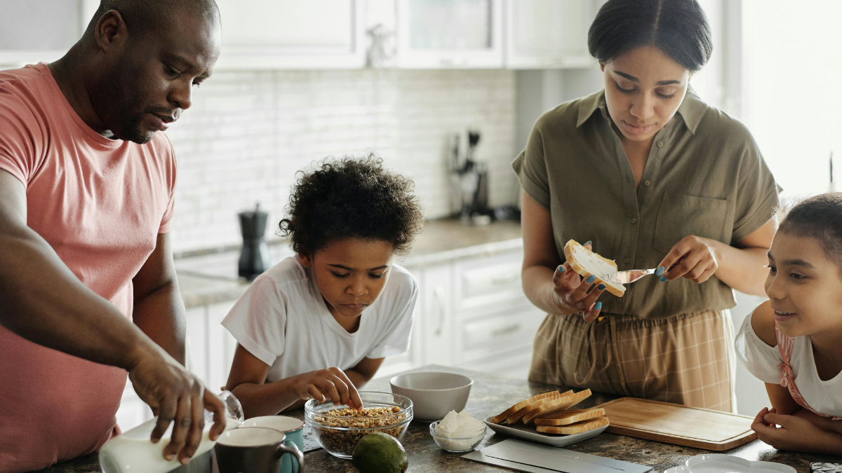Família reunida preparando o café da manhã na cozinha, com alimentos saudáveis como pães, cereais e frutas.